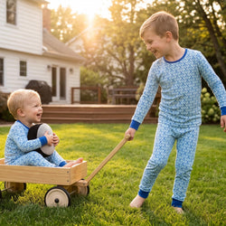 Two young children outdoors on a grassy lawn in front of a house. The older child stands barefoot and pulls a small wooden wagon while wearing light blue hockey patterned pajamas with dark blue trim.