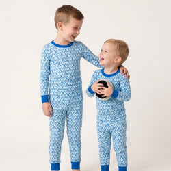 Two young boys wearing matching light blue hockey patterned pajamas with dark blue trim, standing against a plain light beige studio background. The boy on the left has his arm around the boy on the right.