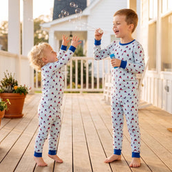 Two young children standing on a wooden porch wearing matching long-sleeve pajamas with a small red and blue hockey player pattern on a white background. One child is blowing bubbles while the other reaches up toward them.