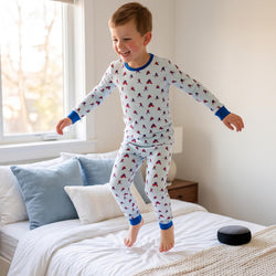 A young boy wearing a white long-sleeve pajama set with a red and blue hockey player pattern jumps on a bed in a bright bedroom.