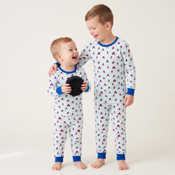 Two young boys standing and smiling, wearing matching long-sleeve pajamas with a white base, blue trim, and a small red-and-blue allover hockey player print. The older boy holds a black plush toy against a plain light beige studio background.
