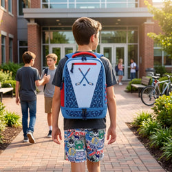 A boy seen from behind wearing a blue and white hockey-themed backpack with crossed hockey sticks printed on the back, walking on a brick campus path toward a building entrance.