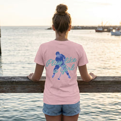 Person wearing a pink t-shirt with a hockey girl graphic and text by a waterfront at sunset