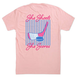Pink t-shirt with hockey graphic and text 'She Shoots, She Scores' on a white background