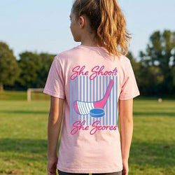 Person wearing a pink t-shirt with 'She Shoots, She Scores' design outdoors.