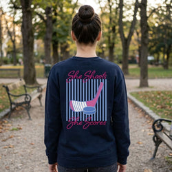 Person wearing a navy sweatshirt with 'She Shoots, She Scores' text and graphics in a park.