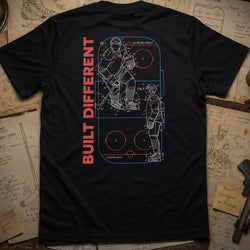 A black short-sleeve T-shirt laid flat on a rustic wooden surface with vintage sports papers and a hockey stick around it. The back of the shirt features a large graphic print with two hockey players outlined in white and red, alongside the words 'BUILT DIFFERENT' in red vertical text.