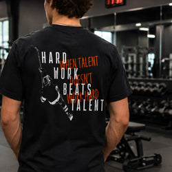 A man viewed from behind wearing a black graphic t-shirt in a gym setting. The back of the shirt features a white hockey player silhouette and the text 'HARD WORK BEATS TALENT' in white, with 'WHEN TALENT DOESN'T WORK HARD' in red.