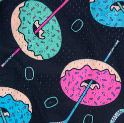 A close-up of a dark navy fabric with a colorful donut print. The pattern features donuts in teal, pink, and blue with light beige edges, decorated with pink, blue, and purple sprinkles, and crossed with diagonal pink and blue hockey sticks.