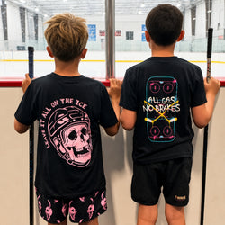 Two boys standing at a window in an indoor ice rink, viewed from behind. Both are wearing black graphic T-shirts and black shorts. One is wearing a Leave It All On The Ice design and the other is in a All Gas No Brakes design
