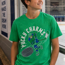 A young boy wearing a bright green graphic t-shirt with a 'Pucky Charms' hockey player design, standing in front of a window looking out onto an ice rink.