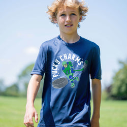 A young boy wearing a navy blue graphic t-shirt with a green and white 'Pucky Charms' hockey player design, standing outdoors on a sunny day with a blurred green field and blue sky in the background.