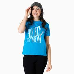 A woman modeling a bright blue short-sleeve graphic t-shirt with 'HOCKEY MOM' printed in white. She is wearing a dark gray baseball cap and black leggings against a clean white background.