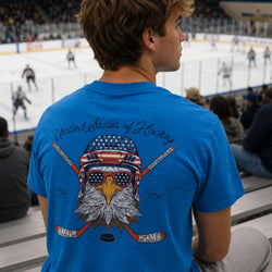 A man viewed from behind wearing a bright blue t-shirt with a large graphic of an eagle wearing an American flag bandana and sunglasses, with crossed hockey sticks and the text 'United States of Hockey' and 'Est. 1776'.