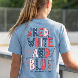 A girl shown from behind wearing a light blue t-shirt with a patriotic red, white, and blue graphic that reads 'RED, WHITE, AND BLUE' with 'ALL AMERICAN HOCKEY GIRL' underneath, set against an outdoor skatepark background.