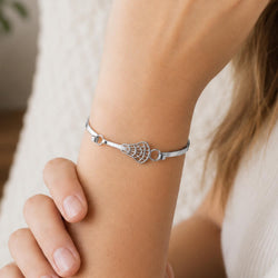 A close-up of a person's wrist wearing a polished silver bangle bracelet featuring a detailed openwork lacrosse head charm.
