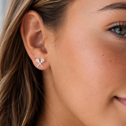A close-up of a woman's ear wearing a small silver stud earring shaped like two crossed lacrosse sticks with clear stones.