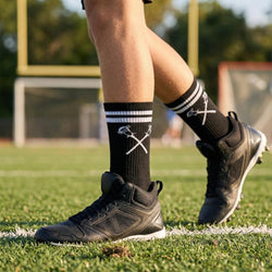 Person wearing black cleats and lacrosse socks on a grass field with a blurred goalpost in the background.