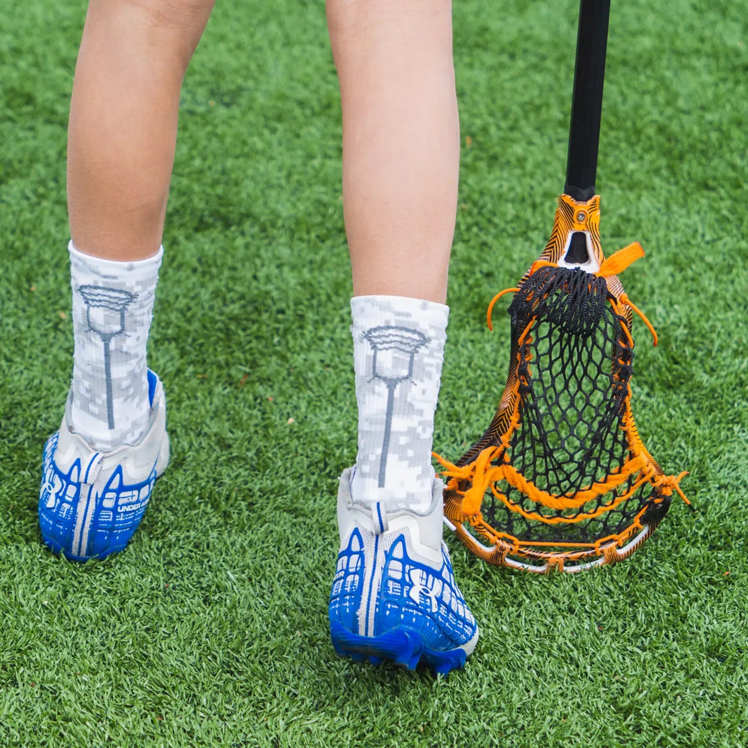 Person wearing lacrosse socks with blue cleats on a grassy field, holding an orange and black lacrosse stick