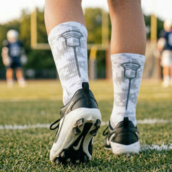 Person wearing cleats and camo socks with lacrosse stick design on a grassy field.