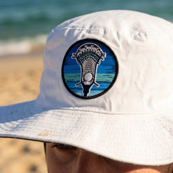 White bucket hat with a blue and black lacrosse emblem on a beach