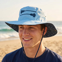 Person wearing a blue bucket hat with a lacrosse stick design on a beach.