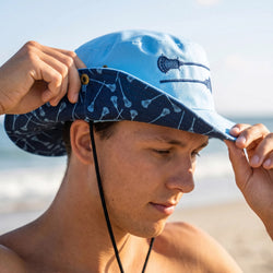 Person wearing a blue bucket hat with a lacrosse stick logo on a beach