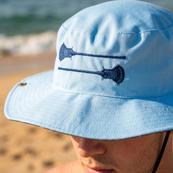 Light blue bucket hat with lacrosse stick design worn by a person on a beach.