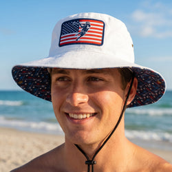 Man wearing a white bucket hat with an American flag design on a beach.