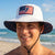 Man wearing a white bucket hat with an American flag design on a beach.