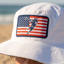 White bucket hat with American flag and lax player design on a beach.