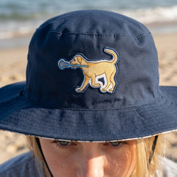 Person wearing a navy bucket hat with a dog and lacrosse stick design on a beach.