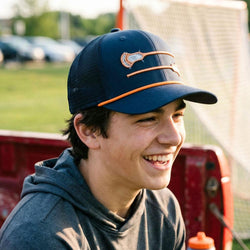 Person wearing a blue cap with orange lacrosse ccents, sitting outdoors near a red vehicle.