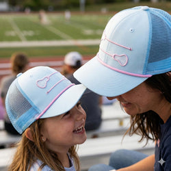 Two people wearing matching light blue caps with pink lacrosse accents on a sports field.