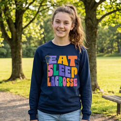 Person wearing a navy long sleeve tee with 'Eat Sleep Lacrosse' in colorful letters, standing in a park.