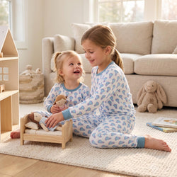Two young girls sitting on a knit rug in a bright living room, wearing matching pajamas with a pink lacrosse stick pattern on a white background. One girl is sitting in a small wooden dollhouse bed holding a stuffed bunny, while the other looks at her with a smile.