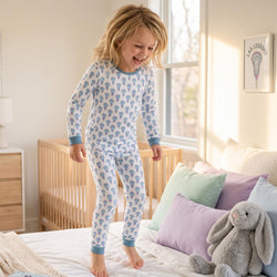 A young child wearing a light blue and white lacrosse patterned long-sleeve pajama set stands barefoot on a bed in a softly lit bedroom. The room includes a wooden crib, pastel pillows, a gray stuffed bunny, and a window with warm sunlight.