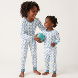 Two children standing barefoot against a plain light background, wearing matching long-sleeve pajamas with a light blue and white lacrosse pattern and blue trim. The older child holds a teal ball.