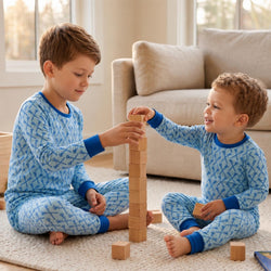 Two young boys sitting on a light rug in a living room, wearing matching blue lacrosse patterned pajamas and stacking wooden blocks together.