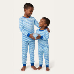 Two children wearing matching light blue lacrosse patterned pajamas with dark blue trim, standing barefoot against a plain light beige studio background.