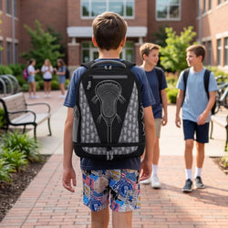 A boy wearing a black and gray backpack with a detailed lacrosse graphic on the back, walking on a campus path with other students in the background.