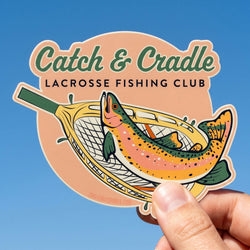 Sticker of a lacrosse fishing club with a fish and lacrosse stick design, held against a blue sky.