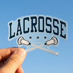 Hand holding a 'Lacrosse' sticker with crossed lacrosse sticks against a clear blue sky.