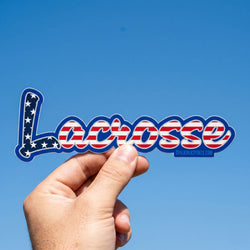 Hand holding a 'Lacrosse' sticker with American flag design against a clear blue sky.