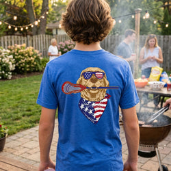 Person wearing a blue t-shirt with a graphic design of a dog holding a lacrosse stick and an American flag bandana, standing in a backyard setting.