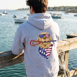 Person wearing a hoodie with a dog graphic and American flag design, standing on a dock by a body of water.