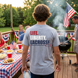 Person wearing a t-shirt with 'LIFE. LIBERTY. LACROSSE' on the back at a casual outdoor gathering.