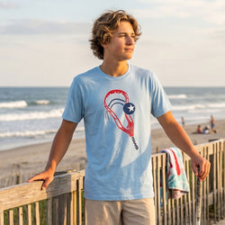 Person wearing a light blue t-shirt with a lacrosse stick graphic design on a beach
