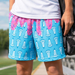 Teen wearing blue shorts with a lacrosse stick pattern and ice cream drip design with a dufflebag on an outdoor field.