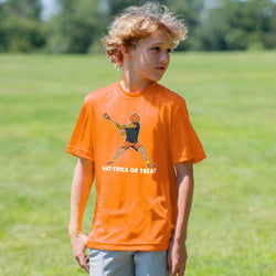 A young boy with curly blond hair stands in a grassy park wearing a bright orange t-shirt with a cartoon pumpkin graphic and the text 'HAT-TRICK OR TREAT'.